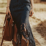 Woman in navy lace midi dress with cowboy boots, brown belt, and leather handbag, outdoor rustic setting